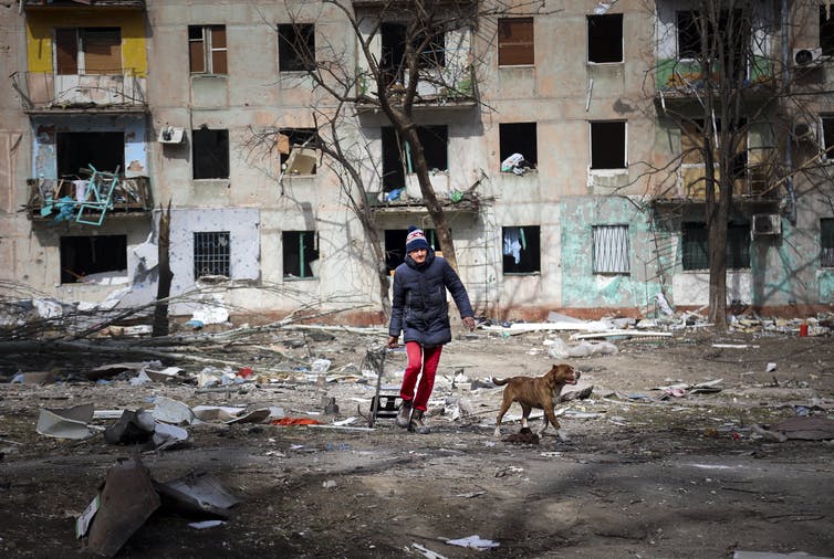 Un hombre con pantalones rojos y una chaqueta azul camina con su perro frente a un edificio de apartamentos destruido.