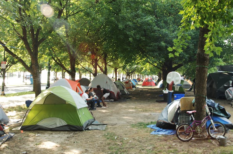 Las tiendas de campaña se alinean en un frondoso parque; se puede ver a algunas personas charlando fuera de una tienda