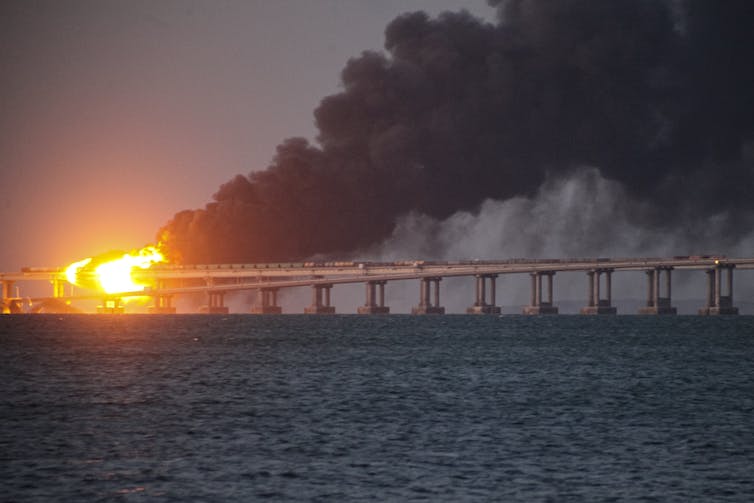 Las llamas y las oleadas de humo negro se arrastran sobre la estructura de un puente sobre un cuerpo de agua.