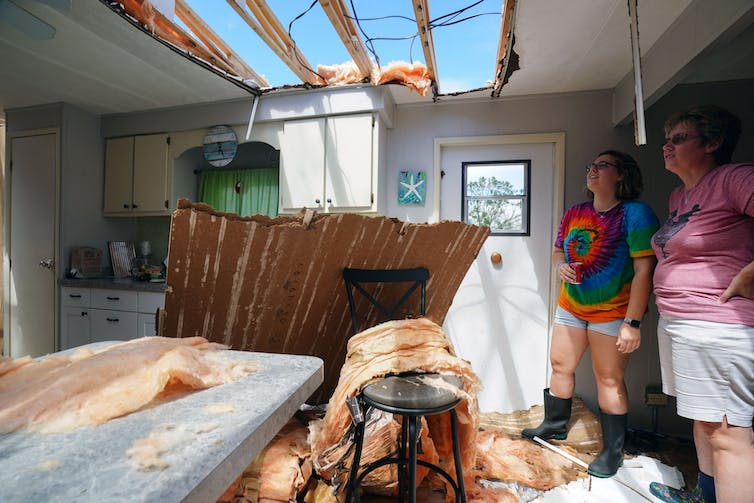 Dos mujeres de pie en una cocina dañada por el viento miran hacia el cielo a través de una sección faltante del techo.