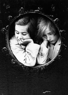 Una foto en blanco y negro muestra a dos niñas mirando a través del ojo de buey de un barco.