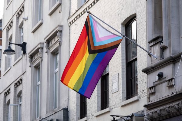 Una bandera con un arco iris, así como rayas negras, marrones, azul claro, rosa claro y blancas en forma de triángulo en el costado, cuelga de un edificio blanco.